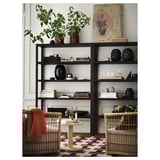 Les élégantes étagères noires STOCKHOLM 2025 contiennent des livres et des objets de décoration, plus grandes que des chaises en osier, avec une plante verte au sommet.