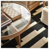 Salon moderne avec table en verre, jeu d’échecs, livres, tapis géométrique, chaise en osier.