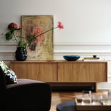 Un salon avec des meubles en bois, une peinture abstraite et un vase en grès noir contenant des fleurs colorées sur une armoire en bois.