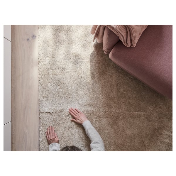 Personne posant un tapis STOENSE beige doux et épais, idéal pour le confort de la chambre et l’isolation phonique.