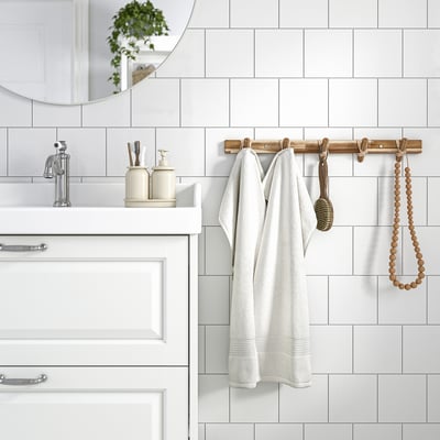 Salle de bain moderne avec porte-crochets en bois STOREDAMM tenant une robe blanche. Tuiles blanches et distributeurs de crème. Bois d’acacia.