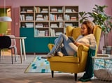 Femme se relaxant dans le fauteuil STRANDMON jaune, en train de lire, entourée d’étagères et de plantes.
