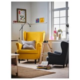 Salon moderne avec grande chaise STRANDMON jaune, oreiller gris, chaise d’appoint noire, échelle en bois, tapis, livres et large fenêtre.