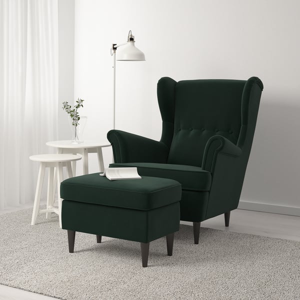 Fauteuil à oreilles STRANDMON moderne en velours vert avec pouf assorti, posé sur un tapis à côté d’une table d’appoint blanche avec une lampe et une plante.