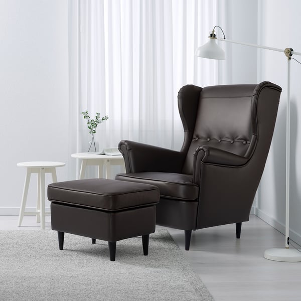 Salon moderne avec fauteuil à oreilles STRANDMON en cuir noir et pouf, table d’appoint blanche avec verdure, tapis gris et lampadaire.