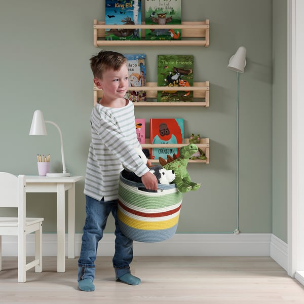 Jeune garçon portant un panier tressé coloré rempli de jouets en peluche, indiquant l’utilisation des STRANDSKATA pour le stockage et la portabilité des jouets.