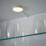 Un projecteur rond moderne monté sur un bras flexible éclaire une étagère en verre avec des verres transparents, démontrant ainsi son utilisation pour l’éclairage des armoires.