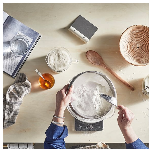 Personne cuisinant, à l’aide d’un tamis d’argent STRÖAD à l’échelle, entourée d’ingrédients et d’outils sur une table en bois.
