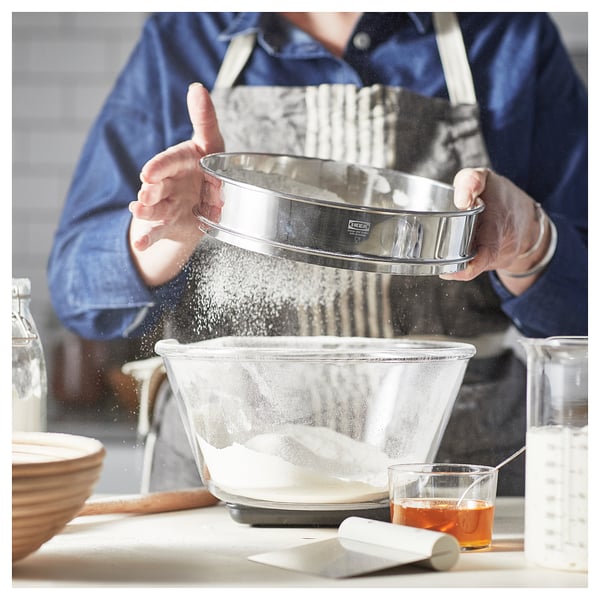 Une personne tamise la farine à l’aide d’un tamis STRÖAD dans un bol, entourée d’outils de cuisson dans une cuisine confortable.
