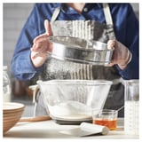 Une personne tamise la farine à l’aide d’un tamis STRÖAD dans un bol, entourée d’outils de cuisson dans une cuisine confortable.
