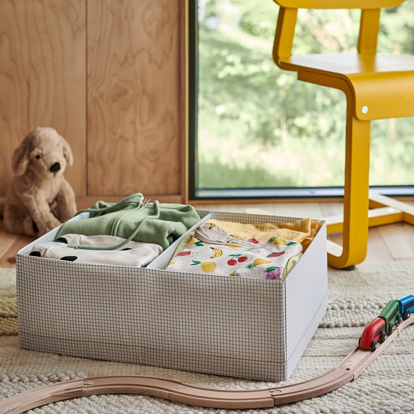 Boîte de rangement ouverte avec les vêtements pliés à l’intérieur. À proximité, un ours en peluche, un train jouet et une chaise jaune sont visibles.