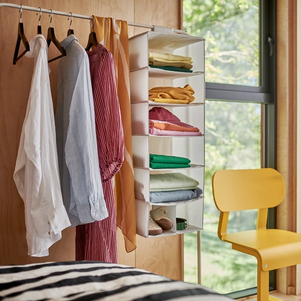 Une scène de chambre à coucher avec un drap de lit rayé, deux cintres contenant des vêtements et une chaise jaune à côté d’une fenêtre.