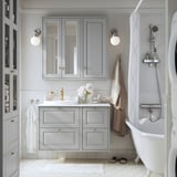 Salle de bains moderne avec meuble TÄNNFORSEN gris clair, section miroir, accents dorés et baignoire îlot blanche. Conception murale peu profonde.