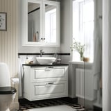 Un meuble-lavabo TÄNNFORSEN en blanc avec un plan de travail noir, des bords classiques, un miroir et des fleurs.