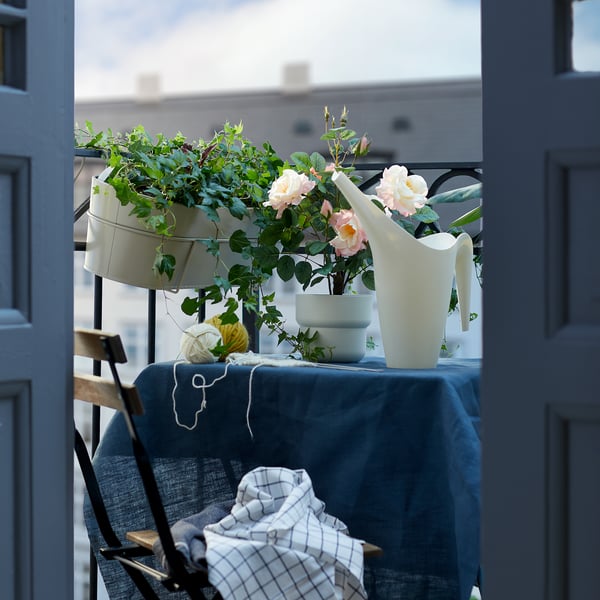 Scène du balcon : jardinières en bois clair avec des fleurs roses, arrosoir blanc, nappe bleue, chaise en métal noir avec une couverture à carreaux.