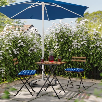 Ensemble de patio extérieur avec table bleue, chaises et coussins sous un parasol bleu, entouré de fleurs blanches.