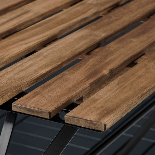 Gros plan d’une table d’extérieur en bois, en acacia naturel avec une teinte semi-transparente, soutenue par des cadres métalliques.