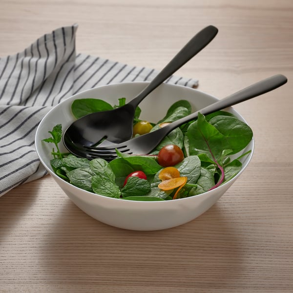Bol blanc de salade verte avec cuillère noire et fourchette, placé sur une table en bois.