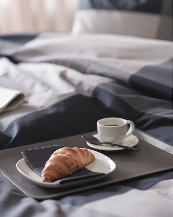 Un plateau TILLGÅNG en plastique polypropylène noir contenant une pâtisserie et une tasse à café sur un lit.