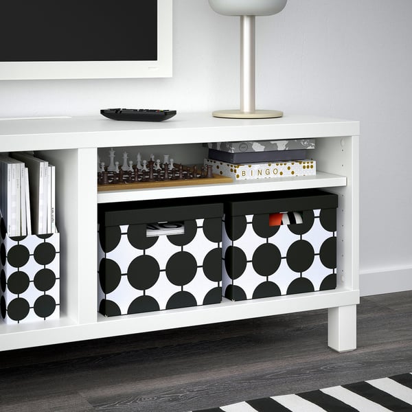 Meuble TV blanc avec étagères de rangement. Comprend des boîtes à motifs noirs et blancs, contenant des jeux comme les échecs et le bingo, des livres et d’autres articles.