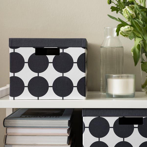 Une boîte TJENA avec des points se trouve sur une étagère, à côté de livres, d’un vase et d’une bougie. Motif noir et blanc, forme rectangulaire, matériau papier.