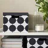 Une boîte TJENA avec des points se trouve sur une étagère, à côté de livres, d’un vase et d’une bougie. Motif noir et blanc, forme rectangulaire, matériau papier.
