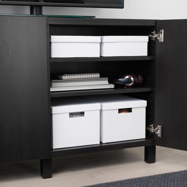 Une armoire noire avec des portes ouvertes révèle un rangement organisé : deux boîtes TJENA blanches, des cahiers empilés et des écouteurs.