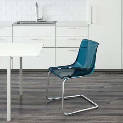 Chaise transparente bleue avec base métallique près de la table de cuisine blanche et de l’évier.