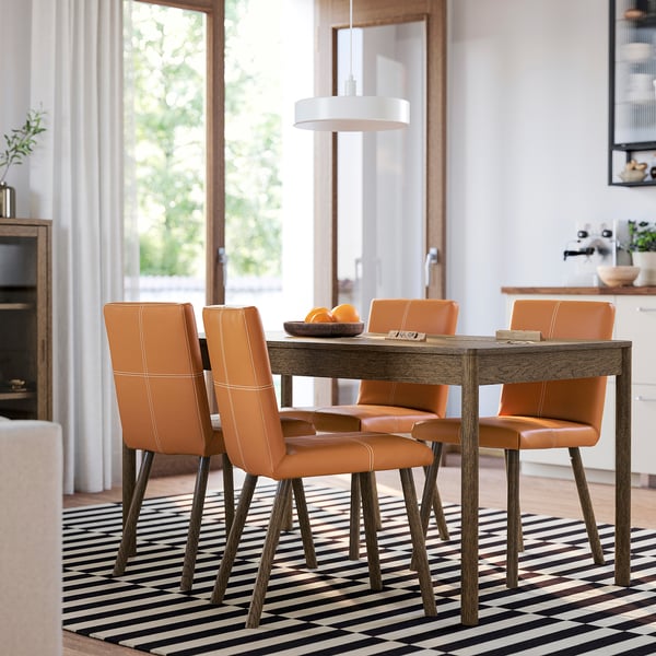 Ensemble de salle à manger moderne : des chaises en similicuir orange entourent la table en bois, sous une grande fenêtre, sur un tapis rayé. Design élégant, petit par rapport à la pièce.