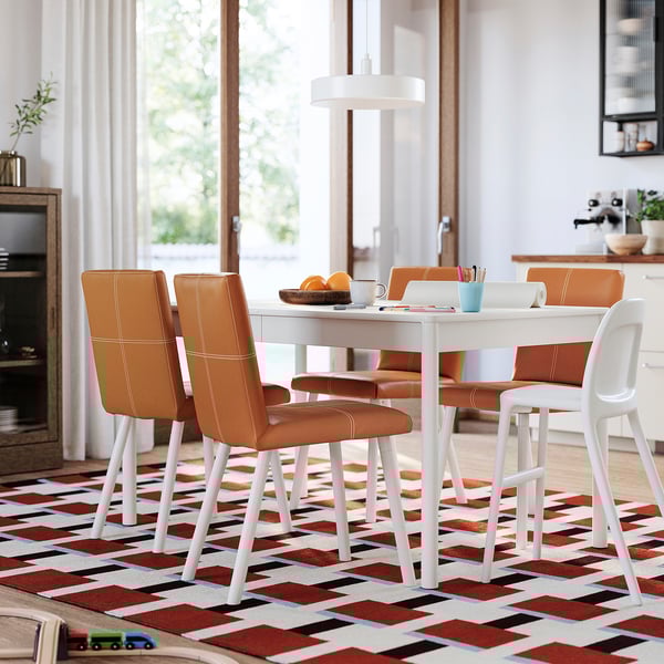 Ensemble de salle à manger moderne avec des chaises en cuir orange et une table blanche sur un tapis coloré dans une pièce bien éclairée.