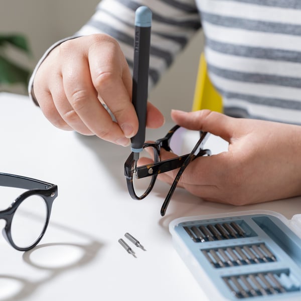 Une personne utilise un tournevis de précision de l’ensemble TRIXIG pour fixer des lunettes. La boîte à embouts magnétique et la poignée sont visibles sur la table.
