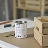 Une petite boîte ronde étiquetée TRIXIG se trouve sur une surface de travail à côté d’un pinceau et d’une caisse en bois non finie.