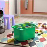 Le bac de rangement vert TROFAST contient des jouets pour enfants sur un tapis coloré, favorisant l’organisation.
