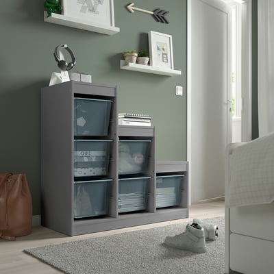 Unité de rangement TROFAST grise avec boîtes en filet, contenant des écouteurs, des sacs et des livres. Il se dresse fièrement, atteignant la hauteur de la porte adjacente.