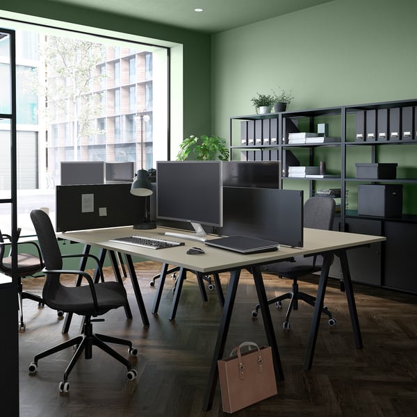 Espace de bureau moderne avec des bureaux trotton en beige, des chaises noires, de grands écrans, un ordinateur portable et des étagères soigneusement organisées contre un mur vert.
