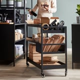 Chariot TROTTEN en métal noir avec plusieurs étagères, contenant diverses fournitures de bureau comme des boîtes en carton, des dossiers et des bouteilles. Facile à déplacer sur roues.