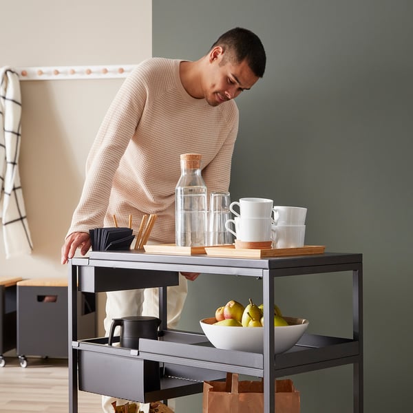 Un homme en pull organise les articles sur un chariot TROTTEN. Le chariot est doté d’une poignée, de roulettes et de compartiments pour les tasses, les fruits et les accessoires.