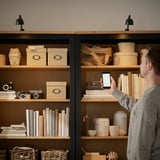 Un homme ajuste son téléphone, éclairant une étagère en bois avec un éclairage d’armoire intégré et réglable, mettant en valeur les objets rangés à l’intérieur.
