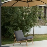 Chaise d’extérieur unique TVARÖ en gris sous un grand parasol beige sur une terrasse, entourée de verdure et faisant partie d’un bâtiment.