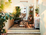 Petit siège d’extérieur avec table et chaises en métal noir, parasol réglable blanc, entouré de plantes, créant un espace ensoleillé confortable.