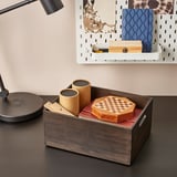 Boîte en bois sur bureau contenant deux récipients cylindriques et un jeu d’échecs. À côté d’une lampe et d’un rangement blanc avec des livres et des objets.