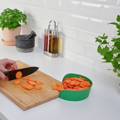 La personne hache des carottes sur une planche de bois, à l’aide d’un bol vert pour recueillir les tranches, sur un comptoir blanc avec des herbes en pot et des distributeurs d’huile.