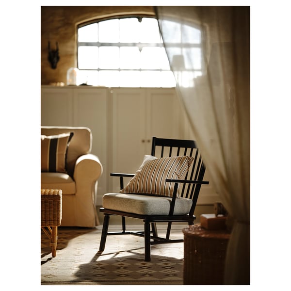 Salon rustique confortable avec fenêtre cintrée, meubles beiges, fauteuil rayé et oreillers, pouf en osier, tapis en laine.