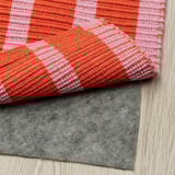 Un tapis à rayures rose et orange vif repose sur un plancher en bois avec une sous-couche antidérapante grise visible en dessous.