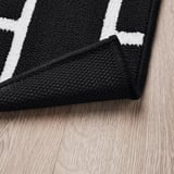 Tapis à carreaux noir et blanc sur parquet. Support antidérapant visible.