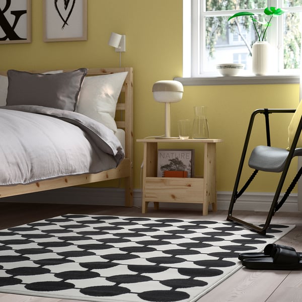 Une chambre moderne avec un tapis VÄNDPLATS noir et blanc, un lit en bois, une literie grise/blanche, un mur jaune et une décoration minimaliste.