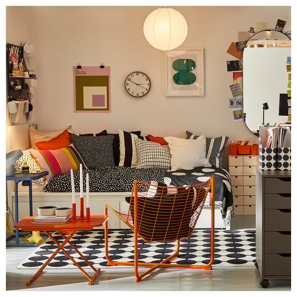 Chambre lumineuse avec tapis à pois noirs, coussins colorés, horloge murale, lampe suspendue, art mural et meubles orange.