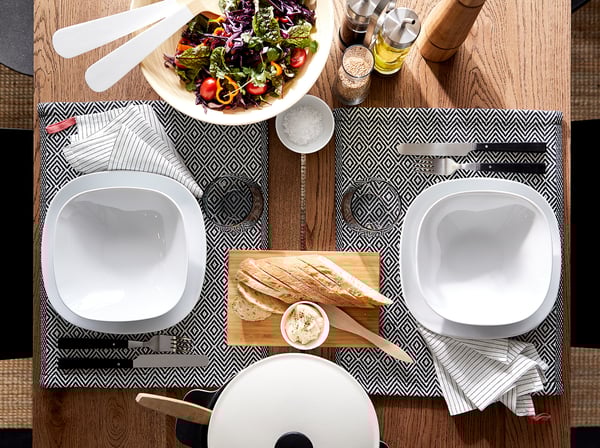 Deux assiettes carrées blanches, un saladier, du pain, des couteaux, des serviettes, sur un chemin de table à motifs.