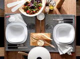 Deux assiettes carrées blanches, un saladier, du pain, des couteaux, des serviettes, sur un chemin de table à motifs.