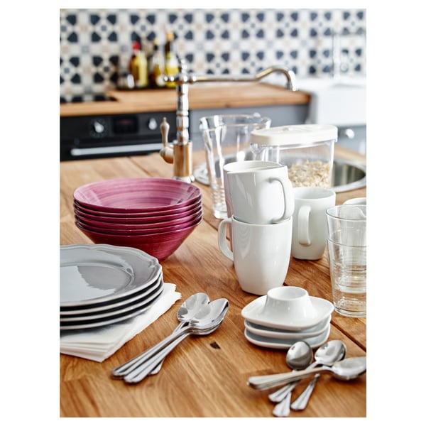 Comptoir de cuisine avec des tasses VÄRDERA blanches empilées, des bols roses, des assiettes grises et des ustensiles en argent contre un mur carrelé.